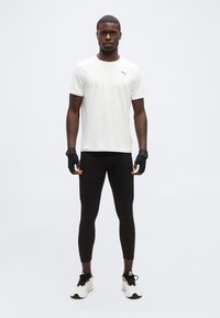 Άνδρας που φοράει λευκό μπλουζάκι Puma, μαύρα κολάν, μαύρα γάντια χωρίς δάχτυλα και λευκά αθλητικά παπούτσια, στέκεται μπροστά σε απλό φόντο.