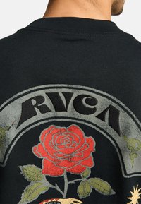 Camiseta negra con un gráfico floral que presenta una rosa roja y hojas verdes, y el texto "RVCA" en letras mayúsculas grises en la parte posterior.