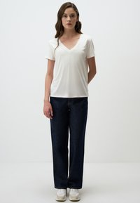 Biała koszulka z dekoltem w serek z miękkiego materiału, zestawiona z ciemnoniebieskimi dżinsami o szerokich nogawkach. Model nosi białe trampki. Prosty, minimalistyczny design.