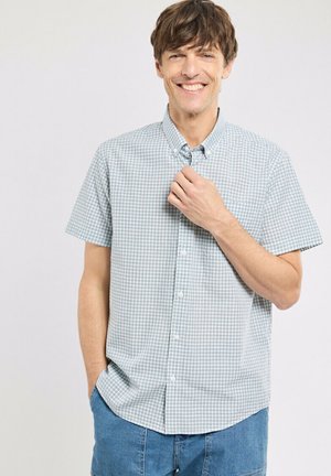 Homme souriant ajustant le col d'une chemise à manches courtes à carreaux vert clair, une main dans la poche, debout devant un fond clair uni.