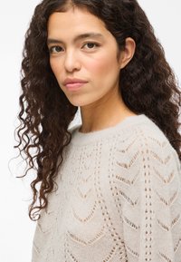 Νεαρή γυναίκα με μακριά σγουρά σκούρα μαλλιά, φοράει ένα κρεμ πανωφόρι από πλεκτό ύφασμα, κοιτάζοντας προς την κάμερα σε έναν λευκό φόντο.