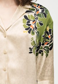 Μπεζ πουκάμισο με κουμπιά και πράσινο, μαύρο και πορτοκαλί λουλουδένιο σχέδιο στον ώμο και το πάνω μέρος του μανικιού, φορεμένο από ένα άτομο με σκούρα σγουρά μαλλιά.