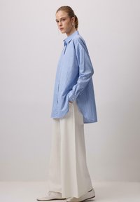 Donna con capelli biondi raccolti in una coda di cavallo, che indossa una camicia a maniche lunghe blu e bianca a righe larghe, pantaloni bianchi larghi e scarpe bianche, in piedi di lato.