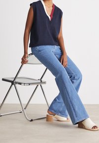 Top senza maniche blu navy con scollatura a V, con dettagli bianchi e rossi, abbinato a jeans svasati azzurri chiari e mule con tacco aperte bianche.