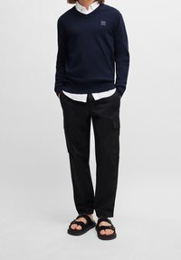 Námořnický svetr s výstřihem do V, černé cargo kalhoty s kapsami a černé sandály s přezkami. Pod tím bílá košile s límečkem.