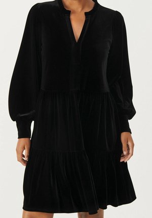Femme portant une robe en velours noir à manches longues avec des volants froncés et un décolleté en V sans col.