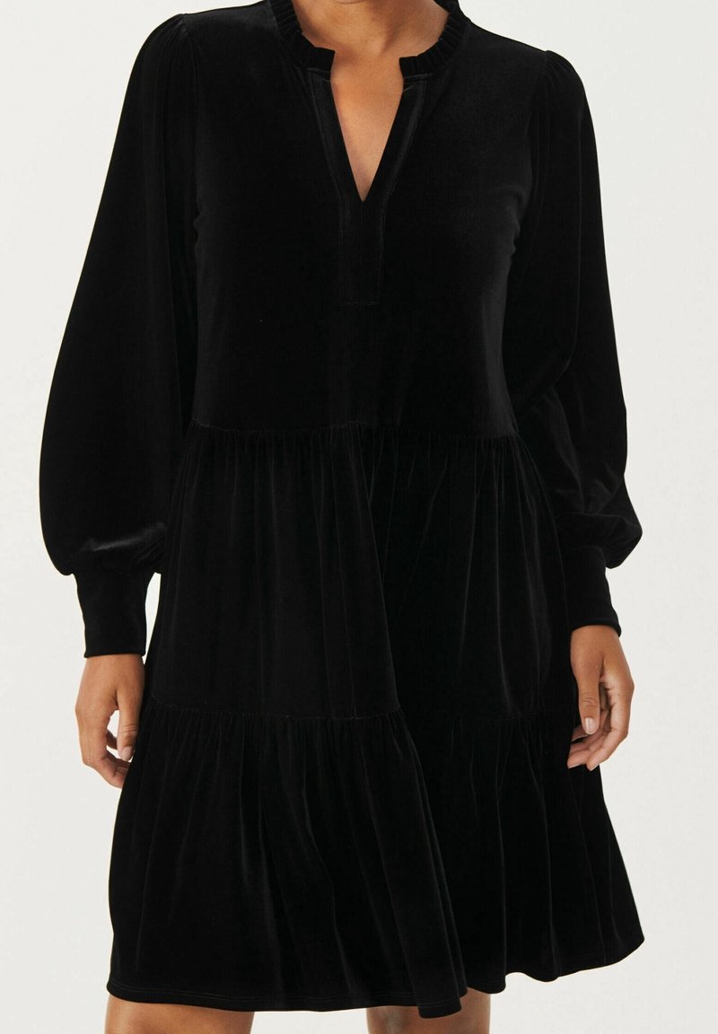 Femme portant une robe en velours noir à manches longues avec des volants froncés et un décolleté en V sans col.