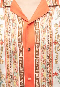 Πουκάμισο με πορτοκαλί γιακά και κάθετες ρίγες που απεικονίζουν floral μοτίβα και επαναλαμβανόμενο κείμενο "Just Cavalli" κατά μήκος της σειράς με τα κουμπιά.