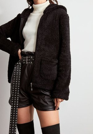 Vrouw draagt een zwarte pluizige jas met ritssluiting, een witte coltrui, zwarte leren shorts met een gespikkelde riem en zwarte kniekousen.