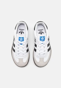 Tênis Adidas com parte superior em couro branco e camurça cinza, riscas pretas e etiqueta do logótipo azul, apresentando uma sola em borracha e cadarços brancos.