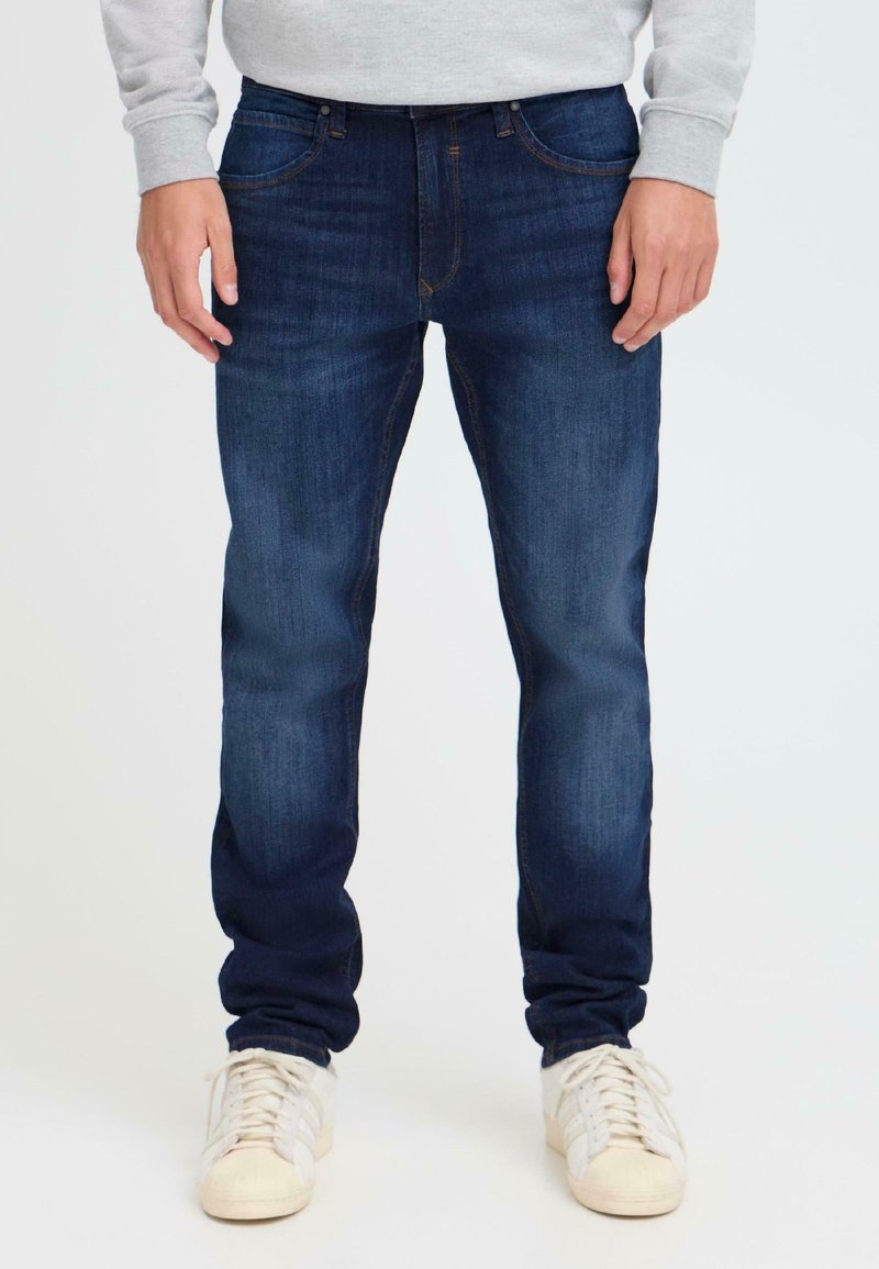 Jeans de mezclilla azul oscuro con corte ajustado, desvanecimiento sutil, dos bolsillos frontales y cierre con cremallera. Llevados con un sudadera gris claro y zapatillas blancas.