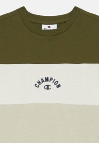 Olive spalvos, baltos ir šviesiai kreminės juostelės medvilninis marškinėliai su apvaliu kaklu ir išsiuvinėtu "Champion" logotipu ant krūtinės.