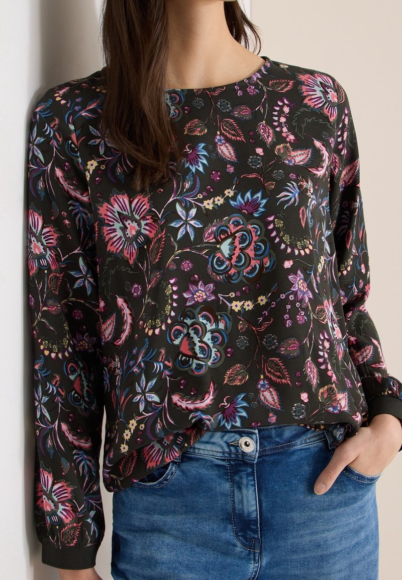 Femme portant un chemisier noir à manches longues avec des motifs floraux colorés et un jean en denim bleu, main dans la poche.