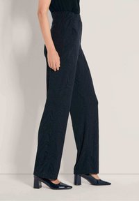 Vrouw in hoge zwarte wijde broek met subtiele textuur en zwarte gepatenteerde leren schoenen met blokhak.