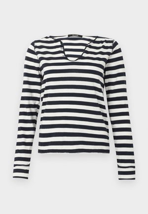 Žena s čiernymi slnečnými okuliarmi, čierno-bielym pruhovaným tričkom s dlhým rukávom, tmavými rozšírenými džínsy a čiernymi špicatými topánkami, stojaca na bielom pozadí.