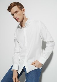 Biała koszula zapinana na guziki wykonana z gładkiej mieszanki bawełny, z klasycznym kołnierzykiem i długimi rękawami. Noszona z niebieskimi dżinsami.