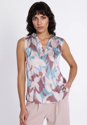 Kobieta o ciemnych włosach do ramion, ubrana w bezrękawnik z kwiatowym wzorem w odcieniach niebieskiego, różowego i mauve oraz jasnoróżowe spodnie, stoi z ręką na biodrze.