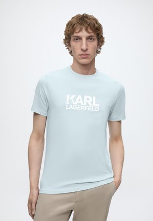 Mlad muškarac s kovrčavom kosom, obučen u svijetloplavu Karl Lagerfeld majicu i bež hlače, stoji ispred jednobojne bijele pozadine.