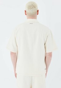 Camisa de manga corta con cuello en color crema claro, fabricada con un tejido texturizado. Detalle de costura notable en las mangas y un pequeño logo en la parte trasera.