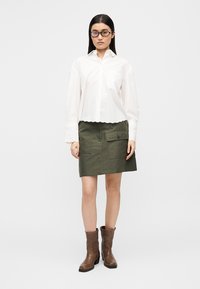 White collared shirt with scalloped hem, paired with an olive green utility skirt featuring side pockets. Brown mid-calf boots complete the look.