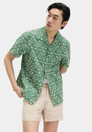 Mann trägt grünes kurzärmeliges Hemd mit Muster und beige Shorts, steht mit verschränkten Händen hinter dem Rücken, schaut seitlich vor einem einfarbigen Hintergrund.