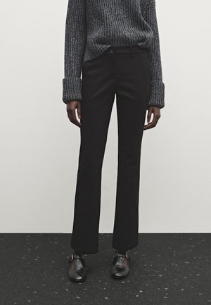 Personne portant un pantalon noir droit, des chaussures noires à boucle et un pull gris côtelé avec les poignets repliés, debout sur un sol sombre.