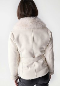 Cremefarbene Jacke mit einem flauschigen Pelzkragen, tailliertem Bund und maßgeschneidertem Design. Weiche Textur, sichtbare Nähte und Nähte auf der Rückseite.