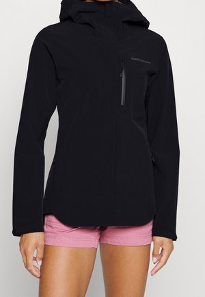 Personne portant une veste à capuche noire avec une poche poitrine zippée et un short rose, debout devant un fond clair uni.