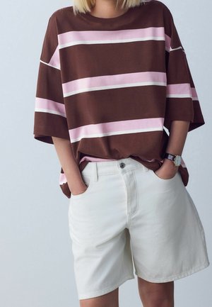 Femme portant une chemise oversize rayée marron et rose avec un short blanc arrivant aux genoux et une montre-bracelet, mains dans les poches.