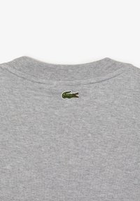 Šedý látkový límec s malým vyšívaným zeleným logem krokodýla umístěným uprostřed pod švem límce.