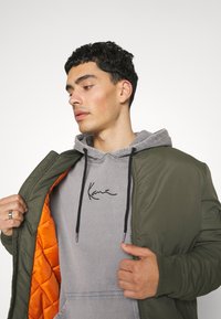 Olivgrüne Bomberjacke mit oranger Innenausstattung, getragen über einem grauen Kapuzenpullover mit schwarzem Schriftlogo. Glattes Material, Reißverschluss, Taschen an den Seiten.