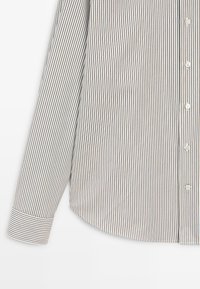 Chemise à boutons rayée blanche, avec des rayures bleu marine et rougeâtres. Elle comporte un col, un poignet réglable avec des rayures similaires, et un ourlet arrondi.