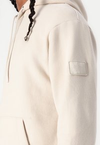 Lys beige hoodie med tekstureret stof, frontlomme med kenguru-lukning, snorhue og et præget logo-patch på venstre ærme.