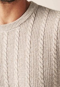 Nahaufnahme eines beigen Kabelstrickpullis mit vertikalen Zopfmustern, der auf der oberen Brust und dem Hals einer Person getragen wird.