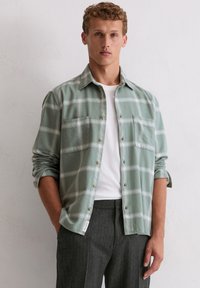 Jonge man in een groen geblokt overhemd met knopen over een wit T-shirt en donkergrijze gestreepte pantalon, staande tegen een effen muur.