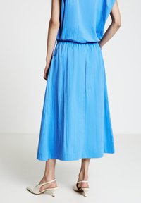 Robe midi bleue en tissu texturé, taille élastique et plis discrets. Associée à des escarpins blancs à bride arrière.