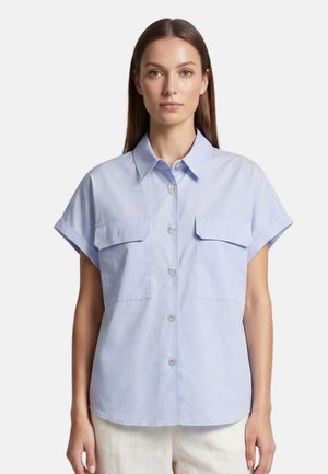 Femme aux cheveux bruns et raides porte une chemise bleu clair à manches courtes avec col, poches avant et boutons, debout devant un fond blanc uni.