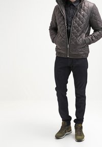 Giacca marrone imbottita con cappuccio, camicia nera con bottoni, jeans scuri e sneakers verde oliva. Presenta una zip frontale e polsini elasticizzati.