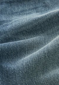 Tela de denim en un tono azul medio, con un tejido texturizado que presenta patrones de espiga visibles y sutiles variaciones en el color.