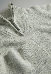 Pilkas megztinis su ketvirčio užtrauktuku, tekstūruota medžiaga ir šonkačio tipo rankogaliais. Jis turi priekinį kišenę, pridedančią detales.