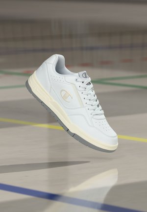 Biela nízka teniska so sivou podrážkou a logom Champion na boku, zobrazená vo vzduchu nad športovou halou s vyznačenými čiarami na podlahe.