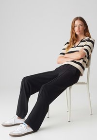 Strickpullover mit schwarz-beigen Streifen, kombiniert mit hochgeschnittenen schwarzen Hosen und weißen Sneakers. Modell sitzt auf einem Stuhl.