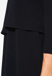 Abito nero a strati con una texture morbida; presenta uno strato superiore fluido sopra una sezione inferiore aderente e dettagli delle cuciture sottili.