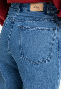 Blå denimjeans med høj talje, en enkelt baglomme med stikninger og et lædermærke ved taljen.