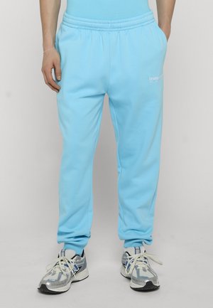 Mann trägt hellblaue Jogginghosen und silber-weiße Turnschuhe, steht mit den Händen in den Taschen vor einem einfarbigen Hintergrund.