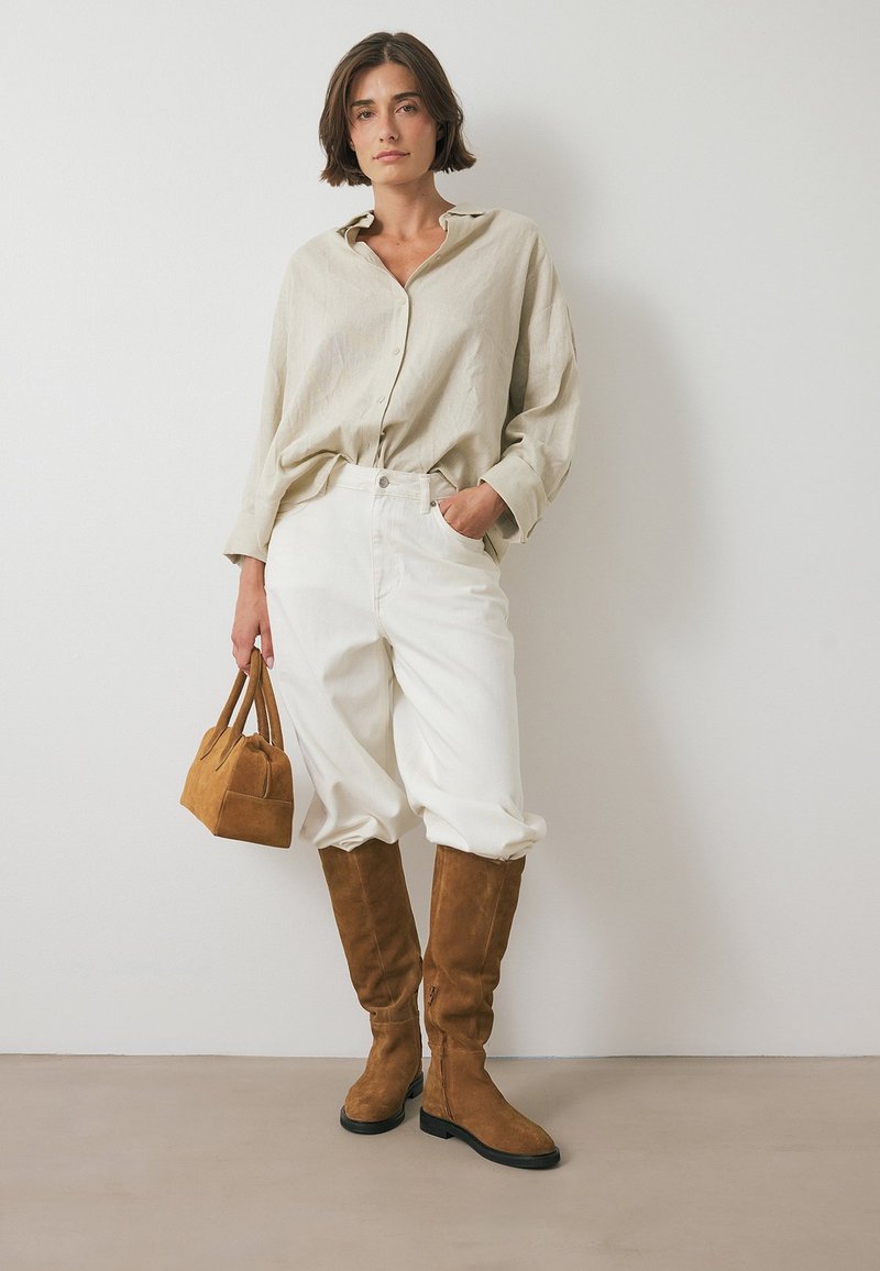 Beige Leinenhemd, weiße übergroße Hose, braune Wildleder-Stiefel bis zum Knie und eine kleine braune Handtasche, vor einem neutralen Hintergrund.