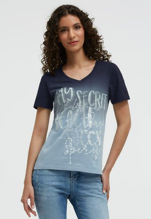 Frau mit lockigem Haar, die ein blaues Ombre-V-Neck-Shirt mit weißem Kursivtext und hellblauen Jeans trägt, steht vor einem einfarbigen Hintergrund.