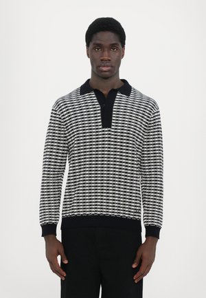 Strickpullover mit einem schwarz-weißen geometrischen Muster, Kragen und gerippten Bündchen. Das Material wirkt weich und strukturiert.