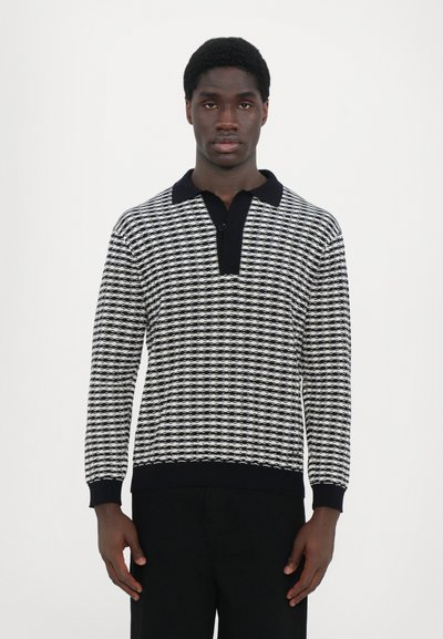 Strikket sweater med et sort-hvidt geometrisk mønster, krave og ribbede manchetter. Materialet virker blødt og struktureret.