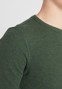 Grön långärmad skjorta med en texturerad, ribbad tyg. Har en rund halsringning och kontrasterande sömnad längs sömmarna.